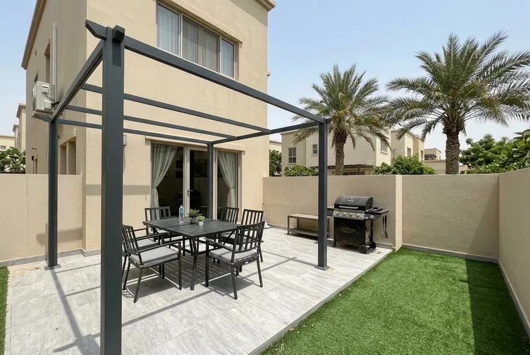 A clean villa patio in Dubai with a dark pergola over light grey tiles, an outdoor dining table, a barbecue and artificial grass